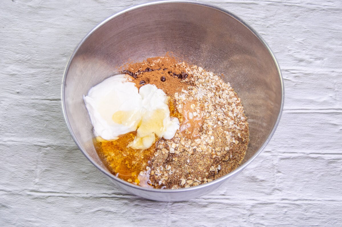 Ingredients for a homemade dog treat recipe in a mixing bowl, including oats, yogurt, banana, and spices, on a white textured surface.