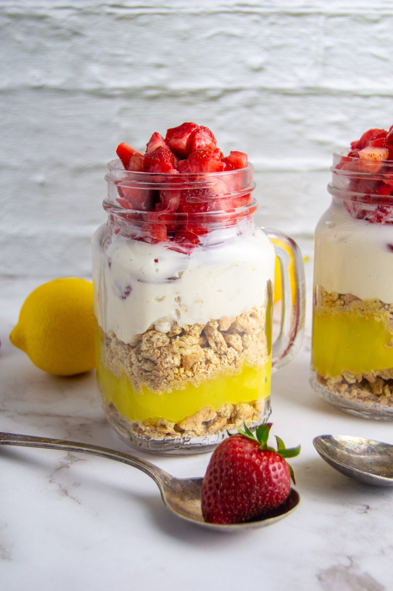 Layered strawberry lemon cheesecake served in mason jars with spoons on the side.