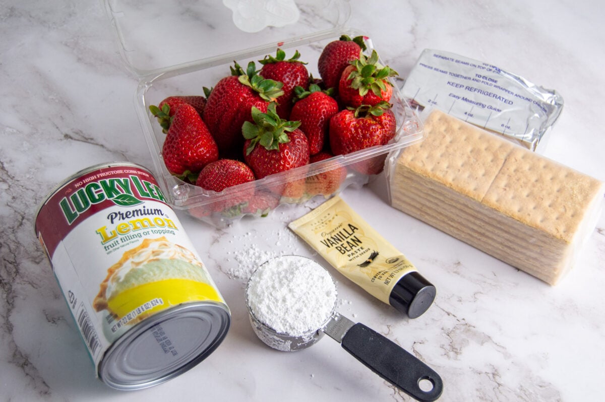 Ingredients for a Strawberry Lemon Cheesecake recipe on a marble countertop, including a can of condensed milk, fresh strawberries, crackers, a packet of cream cheese, vanilla extract, and powdered sugar.