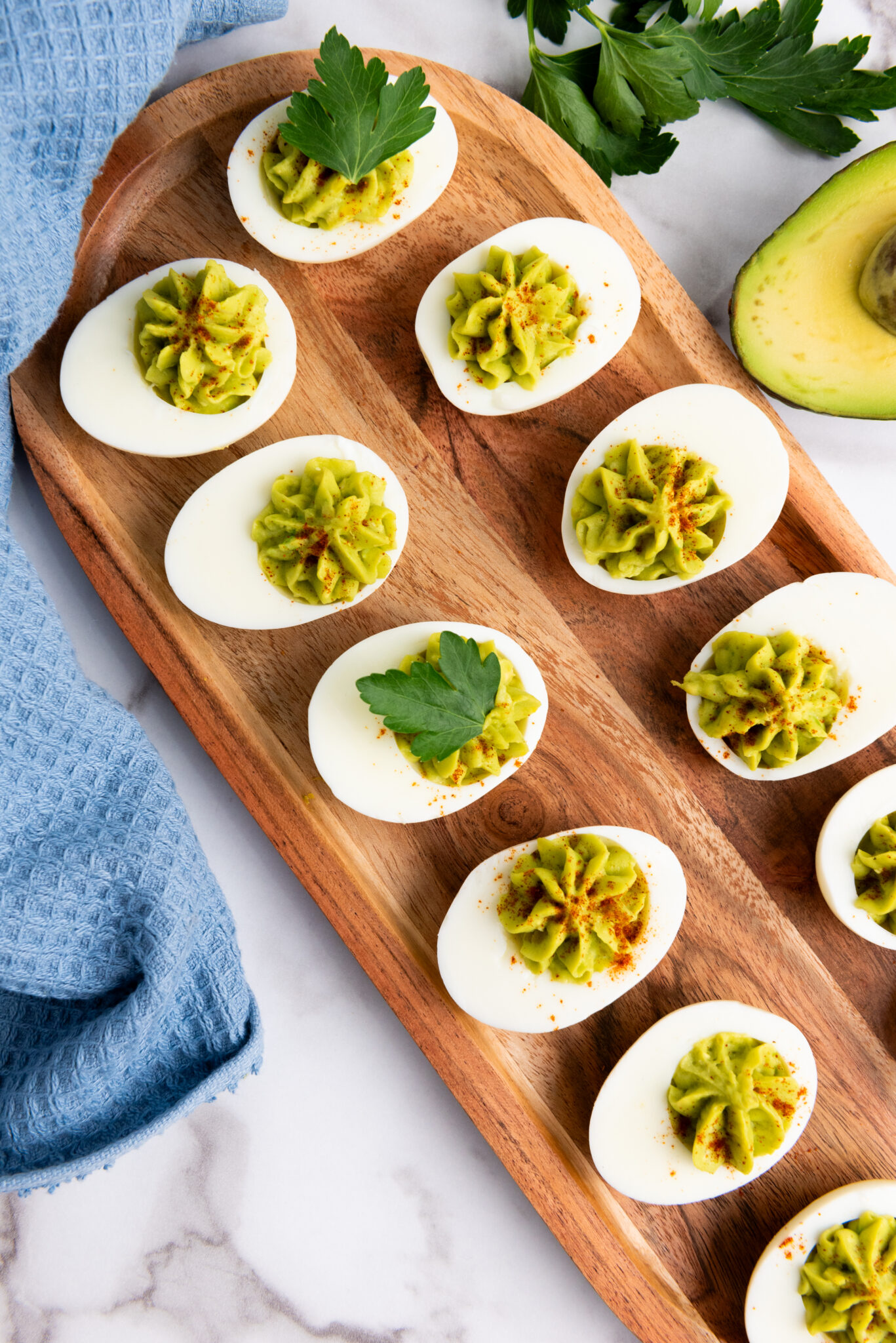 A wooden tray with six deviled eggs filled with a green avocado mixture. The deviled eggs are garnished with fresh parsley and paprika powder.