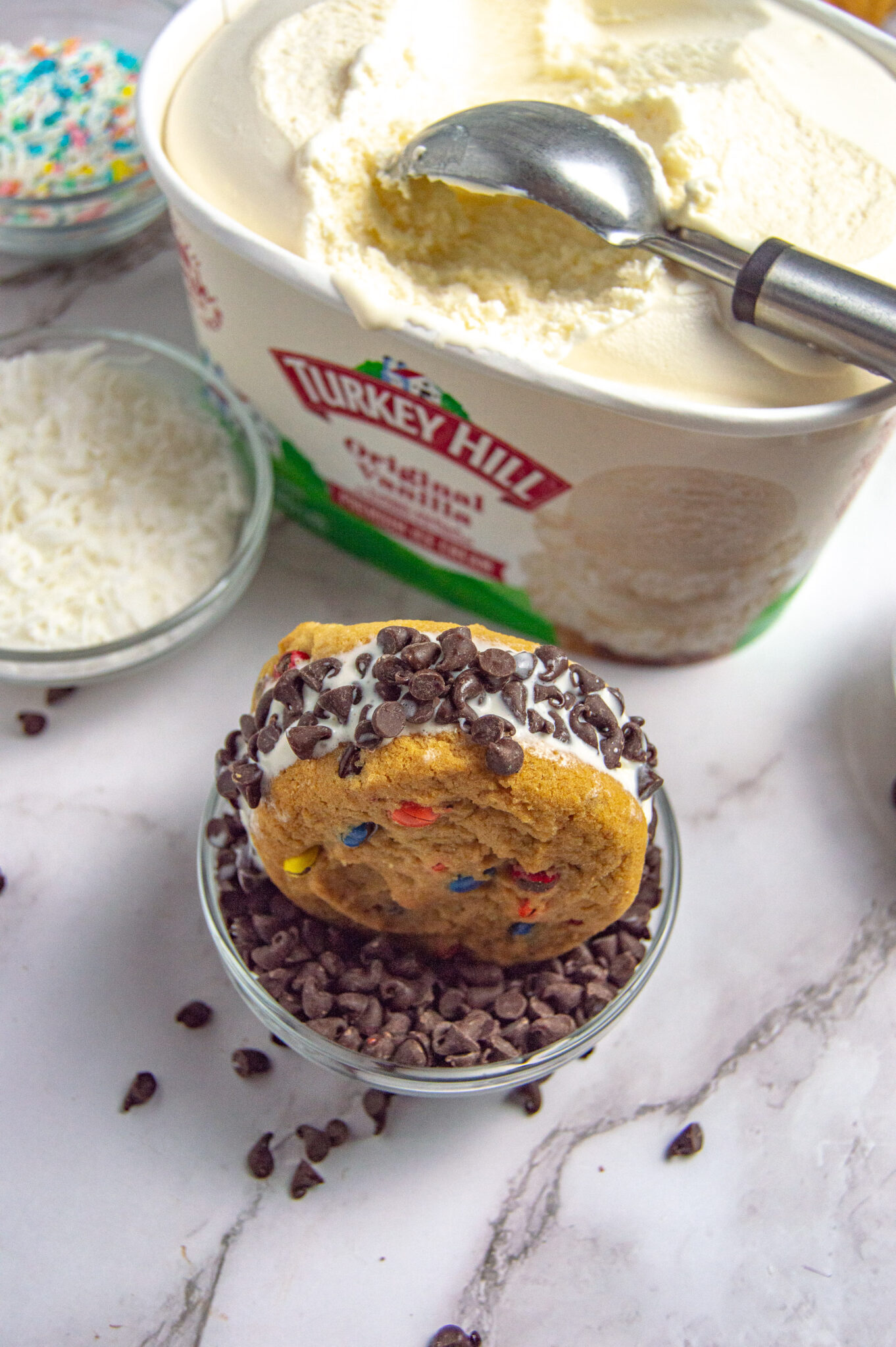 A scoop of classic cookie dough ice cream in a bowl, topped with chocolate chips, with more ice cream and toppings in the background.