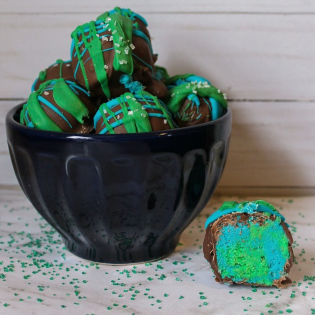 A bowl filled with Earth Day chocolate-covered, bright green cake balls, decorated with drizzled icing and sprinkles, with one cut in half to show a vibrant interior.