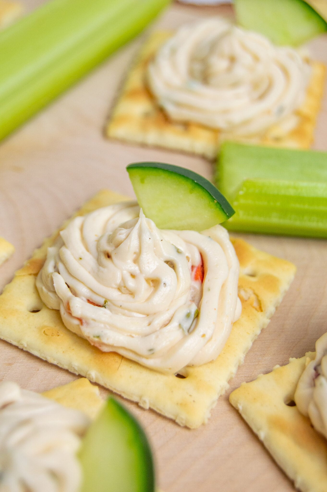 Crackers with a swirl of homemade cream cheese topped with a slice of cucumber, served with celery sticks on a wooden surface.