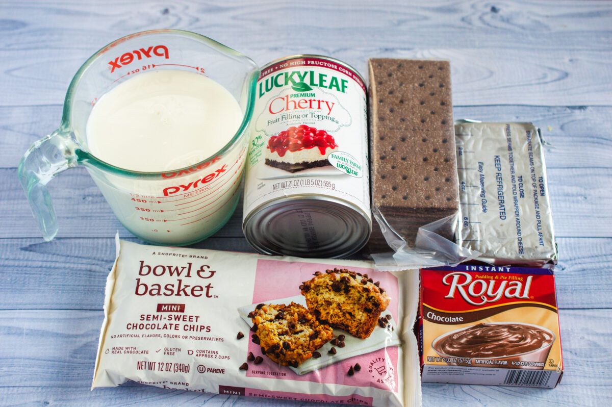 Ingredients for baking on a table, including a measuring cup of milk, chocolate cherry cheesecake filling, chocolate cake mix, chocolate chips, and pudding mix.