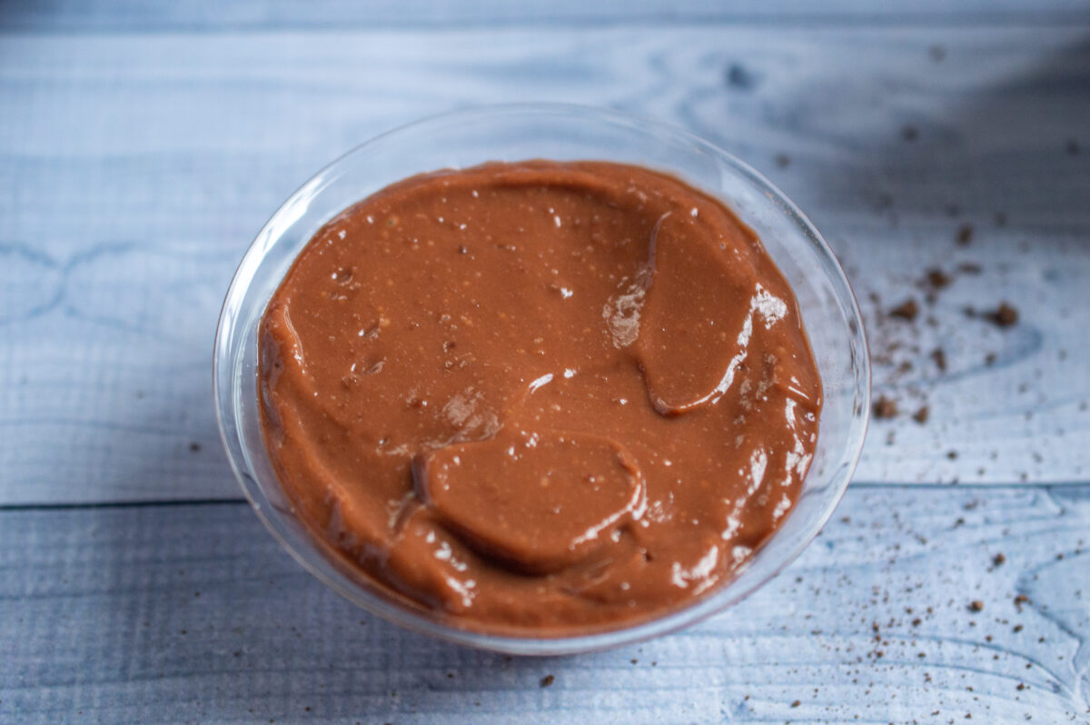 A bowl of smooth, creamy peanut butter on a wooden surface, with a few crumbs scattered around from a no-bake chocolate cherry cheesecake.