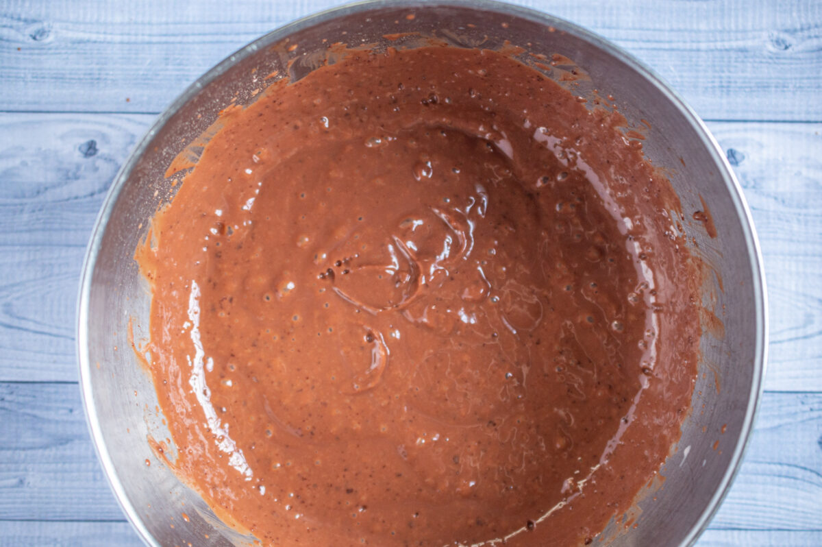 A metal mixing bowl containing mixed chocolate cherry cheesecake batter on a light blue wooden background.
