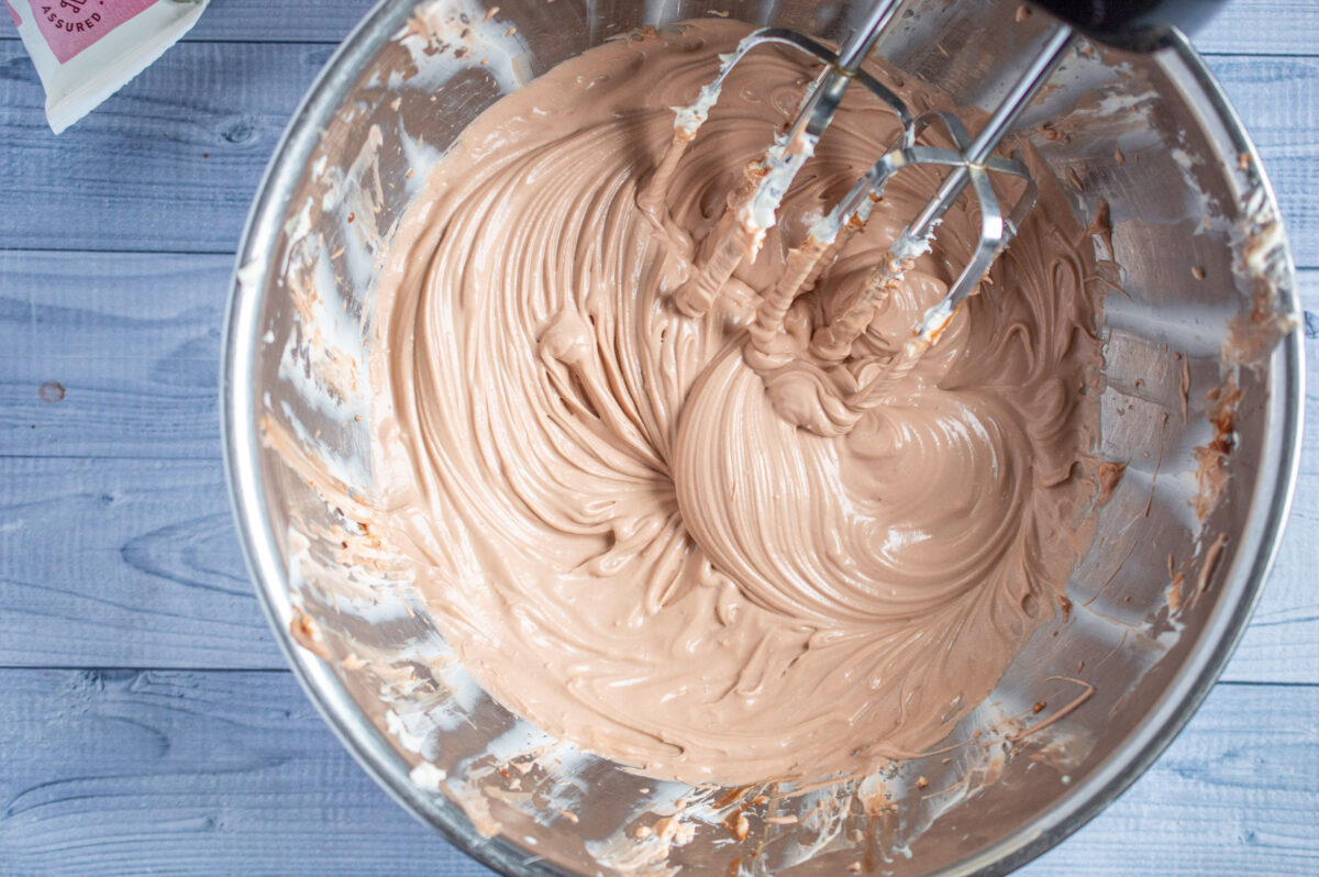 Overhead view of a bowl containing whipped chocolate cherry frosting, with a hand mixer's beaters partially visible, stirring the creamy mixture.