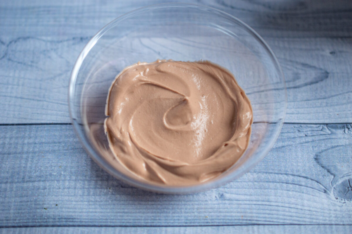 A glass bowl containing smooth, creamy no-bake chocolate cherry cheesecake frosting on a blue wooden surface.