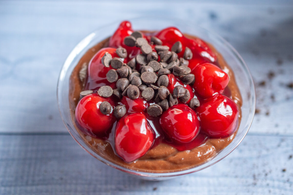 A bowl of chocolate pudding topped with glossy red cherries and chocolate chips, presented on a wooden surface as a preview of the No Bake Chocolate Cherry Cheesecake recipe.
