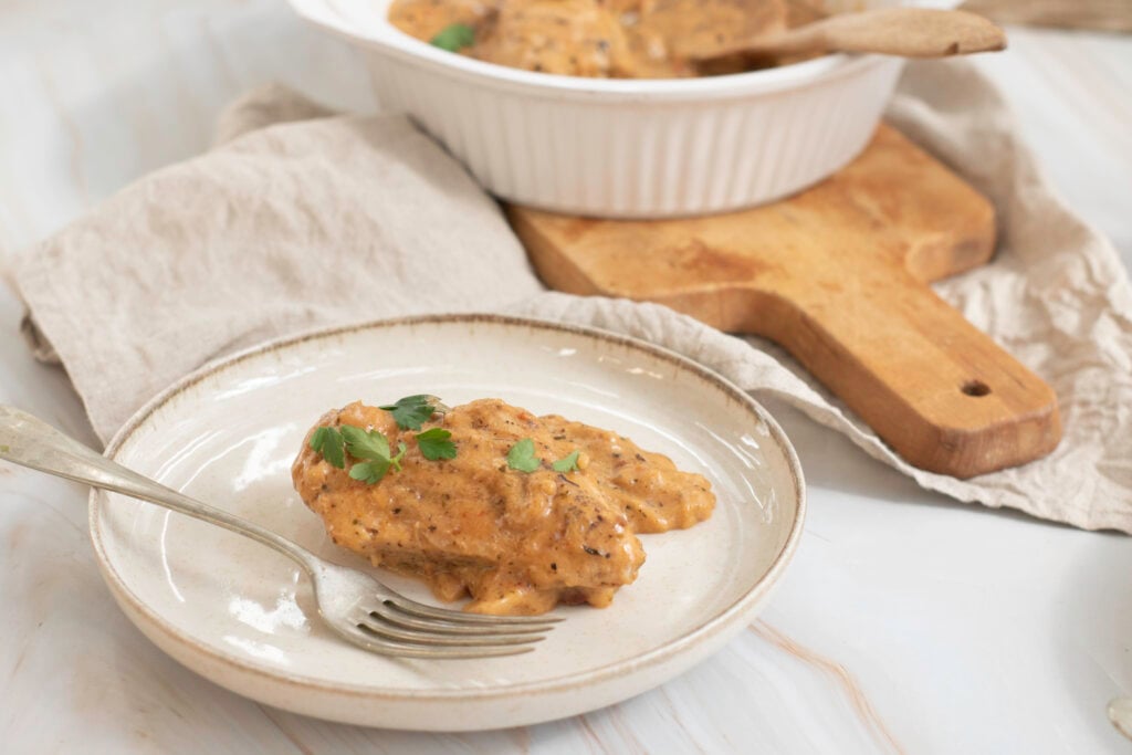 A plate with a piece of marry me chicken topped with a creamy sauce and parsley, alongside a serving bowl with more chicken and a wooden cutting board.