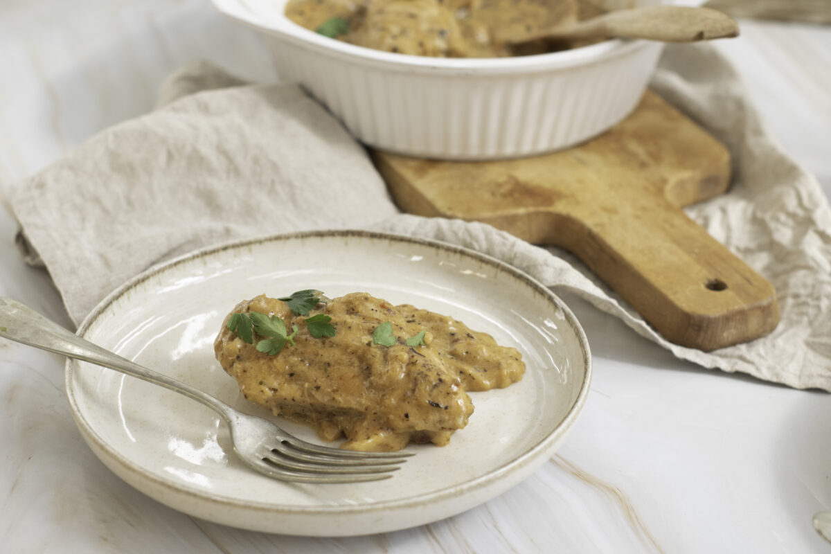 A plate with a piece of marry me chicken topped with a creamy sauce and parsley, alongside a serving bowl with more chicken and a wooden cutting board.