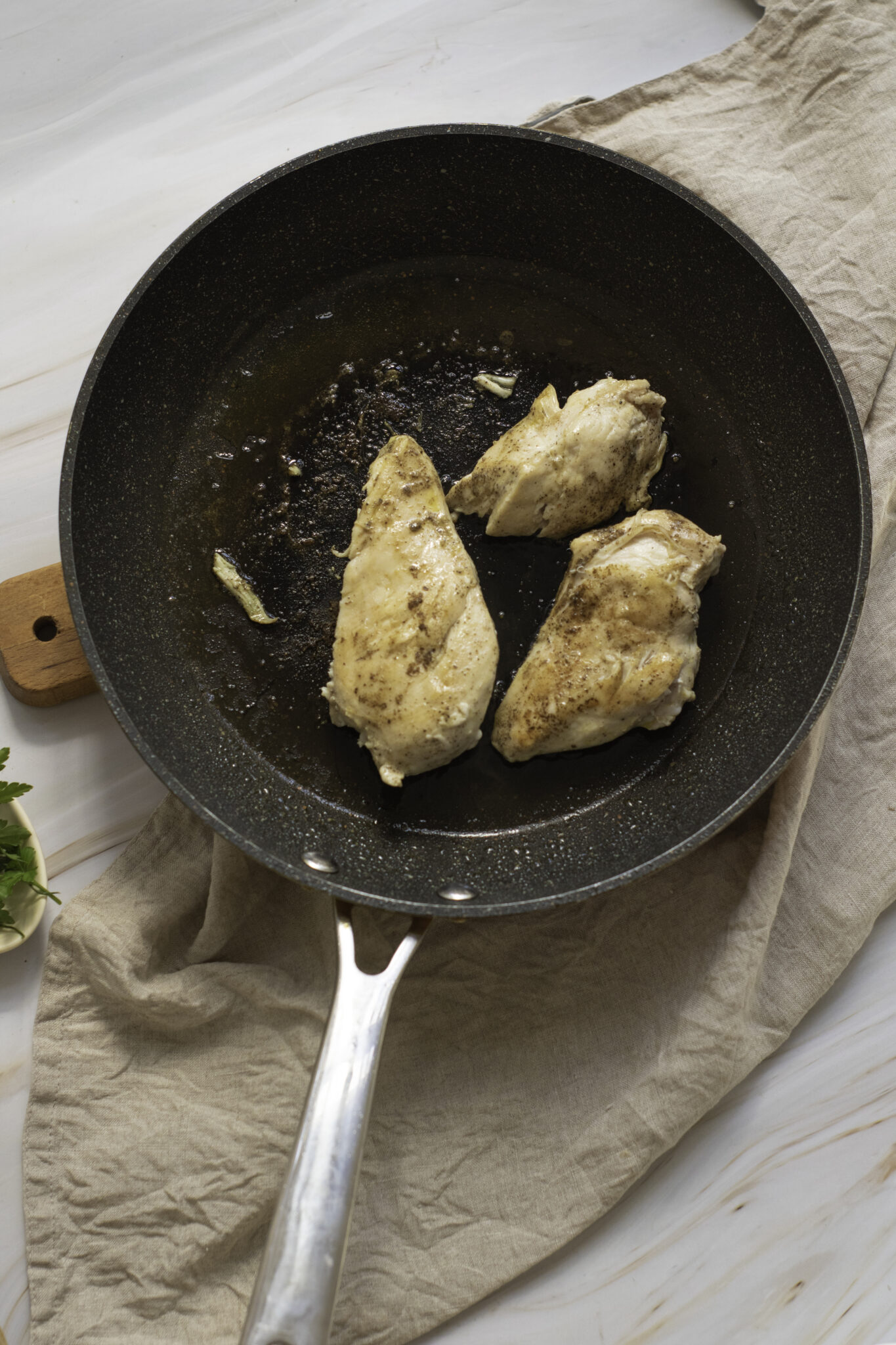 Three pieces of Marry Me Chicken in a black frying pan on a wooden table, covered with a beige cloth.