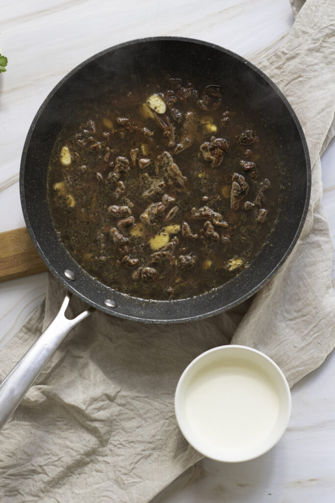 A skillet with simmering beef stew alongside a small bowl of cream on a kitchen counter, part of a "how to make" recipe series.