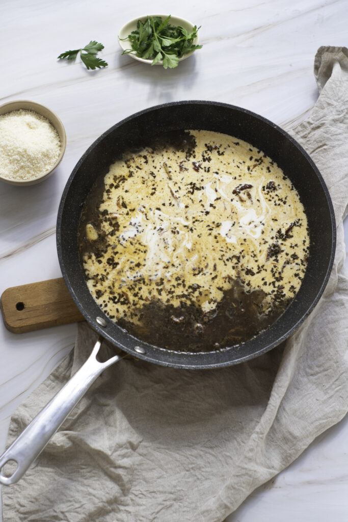 A skillet with simmering creamy sauce and herbs for "marry me chicken," on a kitchen towel beside fresh greens and a bowl of grated cheese.