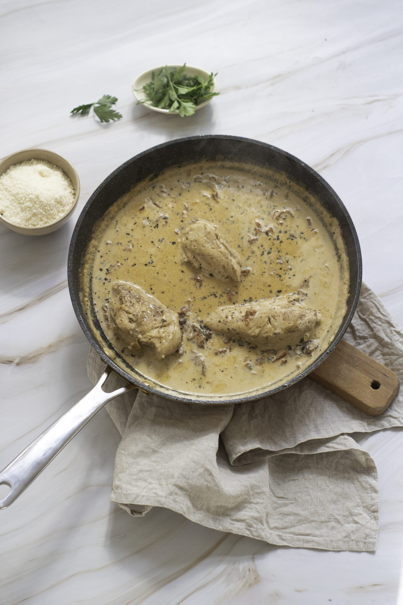 A skillet of Marry Me Chicken on a wooden surface, garnished with herbs, beside a bowl of grated cheese and fresh greens.