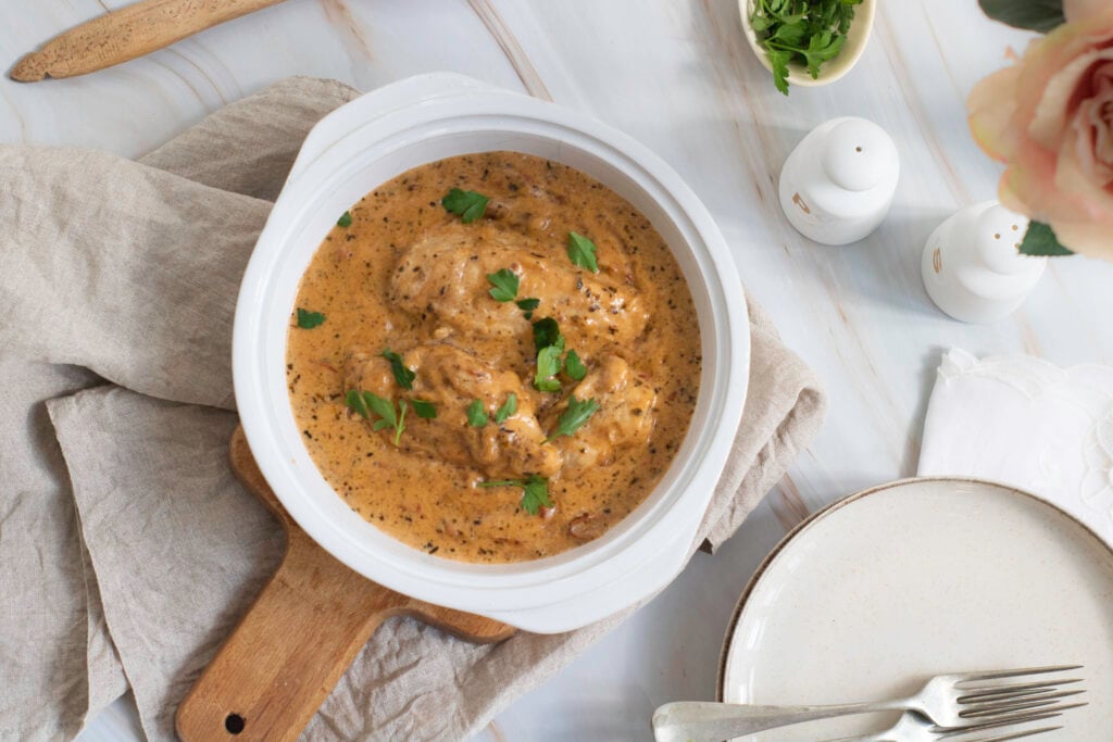 A bowl of creamy marry me chicken garnished with herbs on a wooden board, surrounded by cutlery, a napkin, and a small vase with flowers.