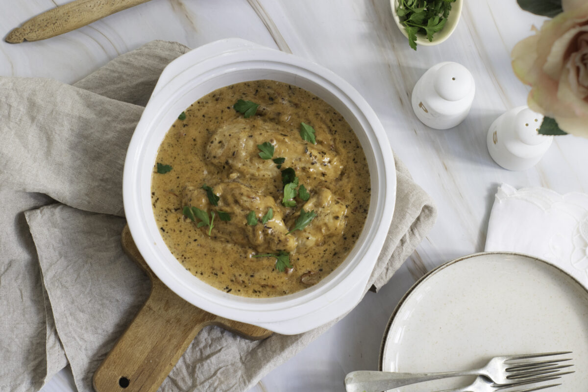 A bowl of creamy marry me chicken garnished with herbs on a wooden board, surrounded by cutlery, a napkin, and a small vase with flowers.