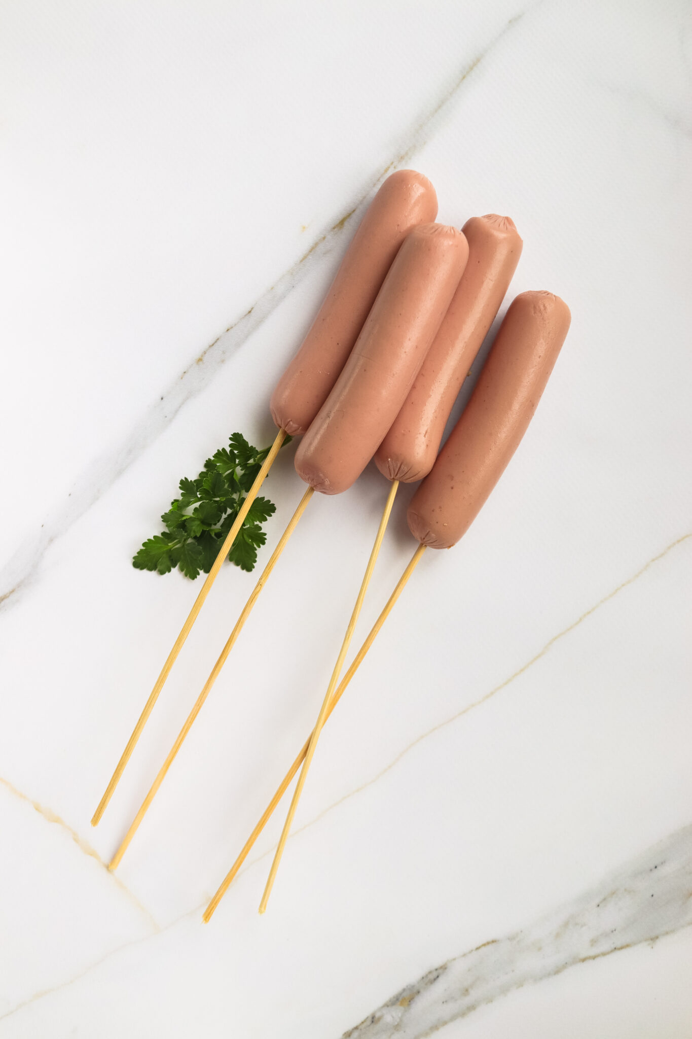 Four hot dogs skewered on sticks are arranged in a row on a white marble surface, resembling a State Fair recipe, and accompanied by a small sprig of cilantro.