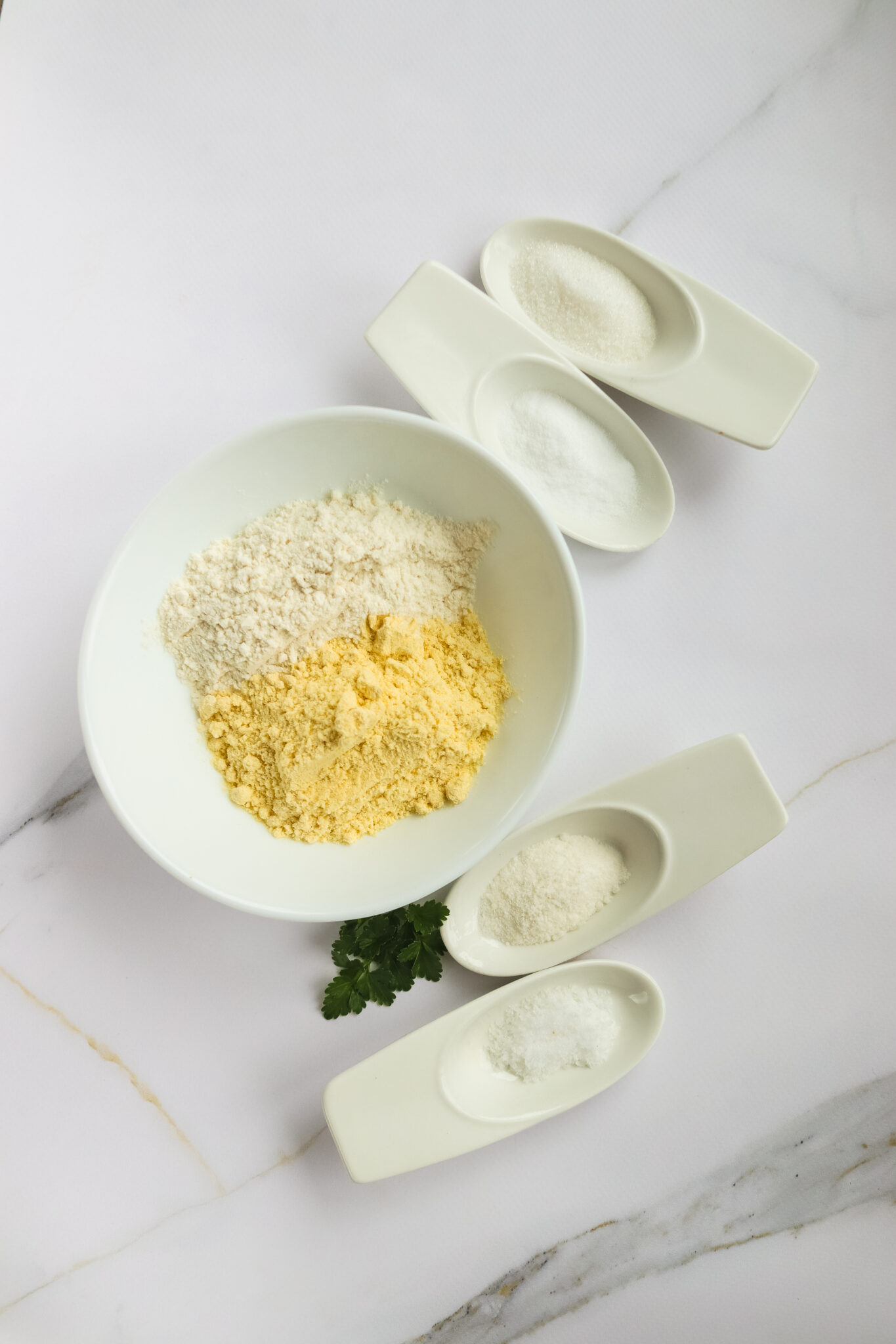 A white bowl with two types of flour and four white oval dishes containing powdered ingredients, on a marble surface. A sprig of green herbs is placed next to the bowl, evoking the charm of a State Fair recipe preparation.