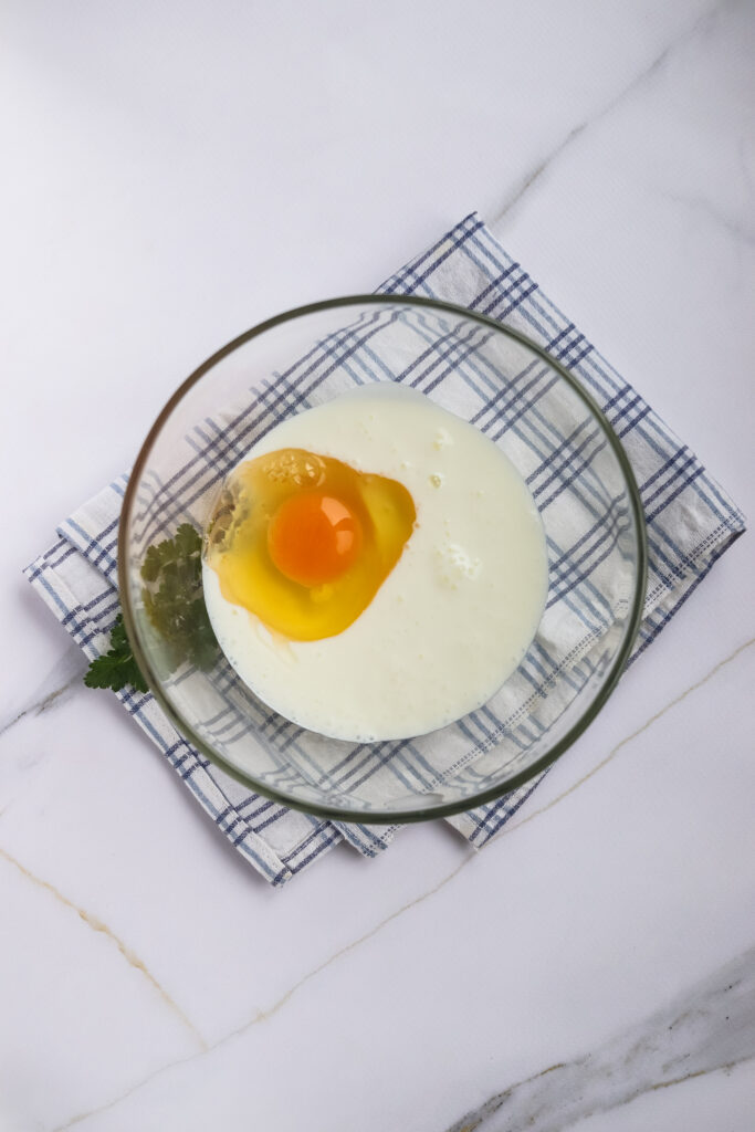 A raw egg is cracked into a glass bowl containing liquid, ready for the perfect corn dog recipe, placed on a blue and white checkered cloth on a white marble surface.