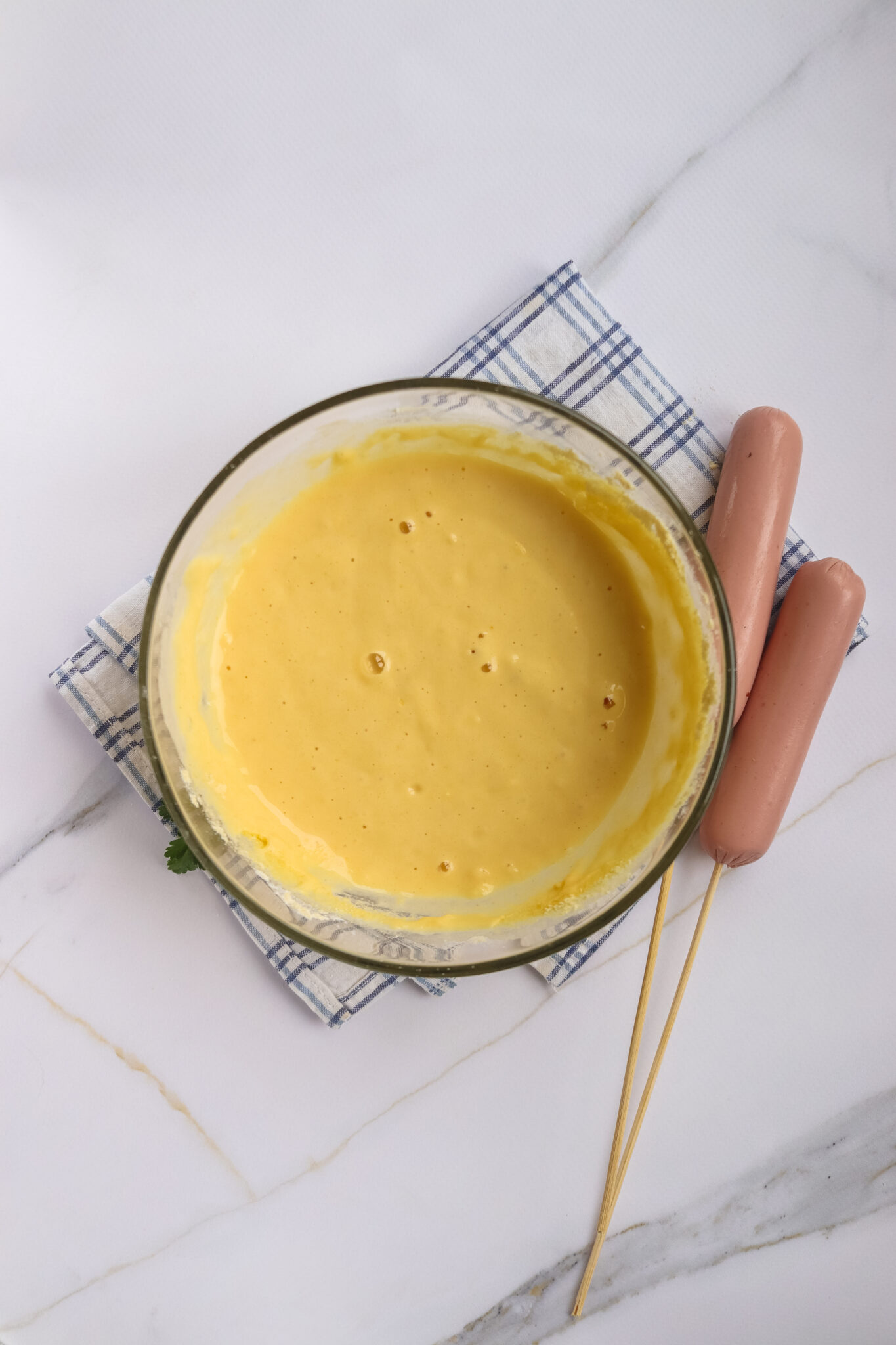 A glass bowl of yellow batter sits on a checkered cloth, ready for the perfect corn dog recipe, with two skewered hot dogs beside it on a white marble surface, evoking the nostalgia of your favorite state fair treats.
