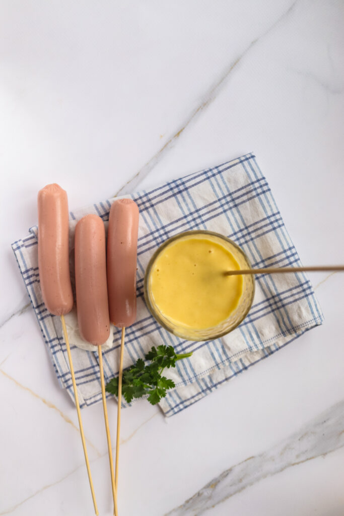 Three skewered sausages next to a small bowl of mustard on top of a blue and white checkered napkin, reminiscent of a State Fair favorite, garnished with parsley.