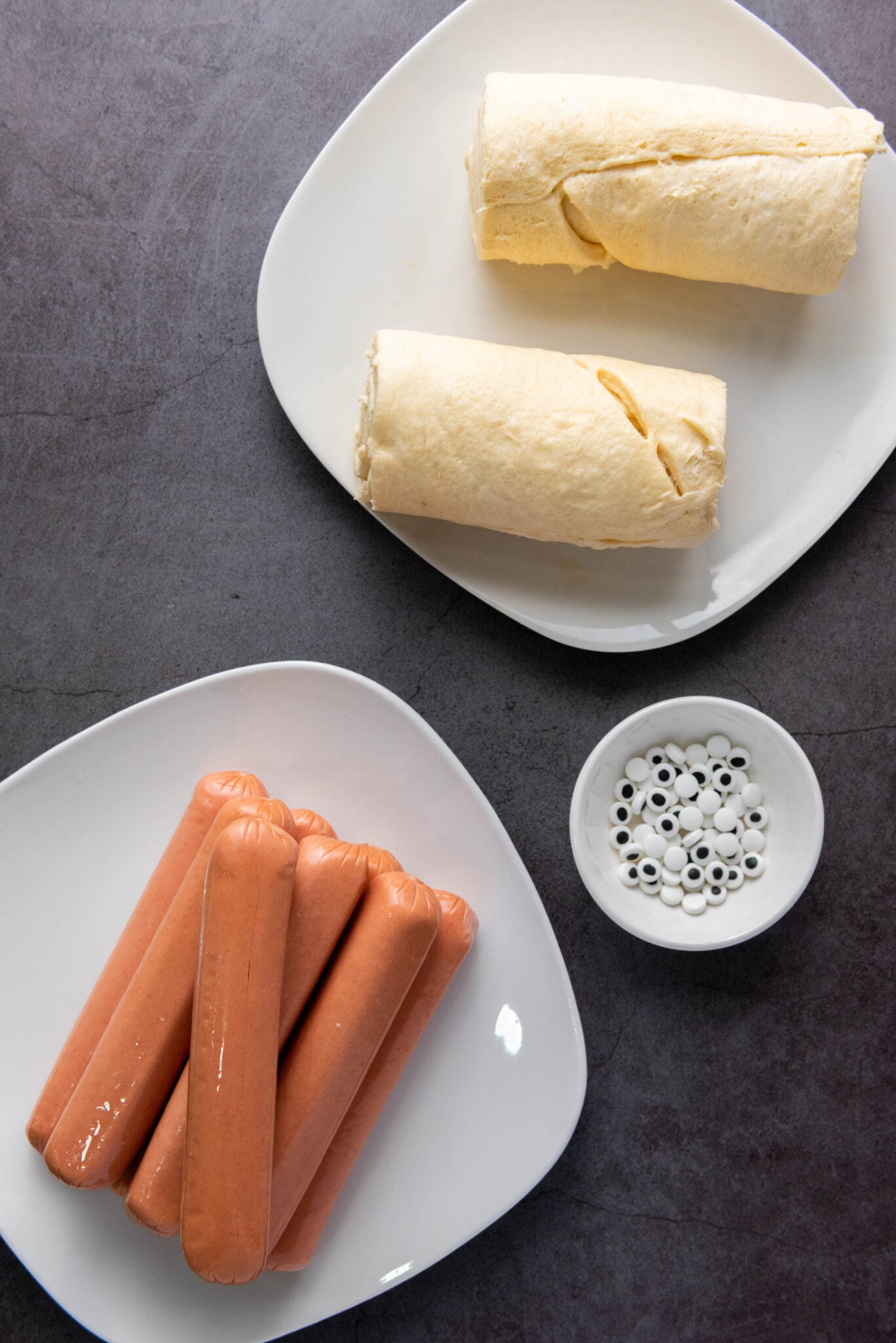 Two plates: one with six uncooked hot dogs, another with two unbaked crescent rolls. Nearby, a small bowl contains googly eyes, adding an element of fun to the auto-draft of your meal plan.