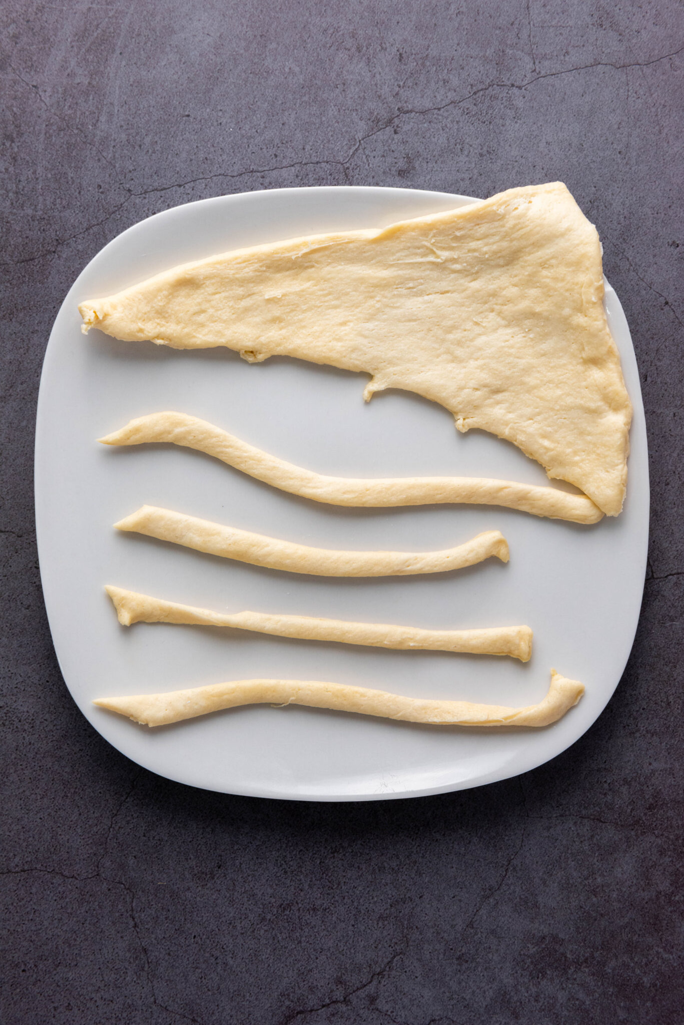 Dough pieces arranged on a white plate; one large triangular piece at the top and four long, thin strips below it. The plate, reminiscent of an Auto Draft layout, is set on a dark, textured surface.