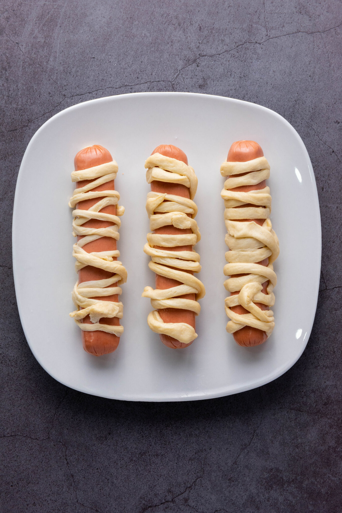 Three hot dogs wrapped in dough arranged in a row on a square white plate, placed on a gray auto draft surface.