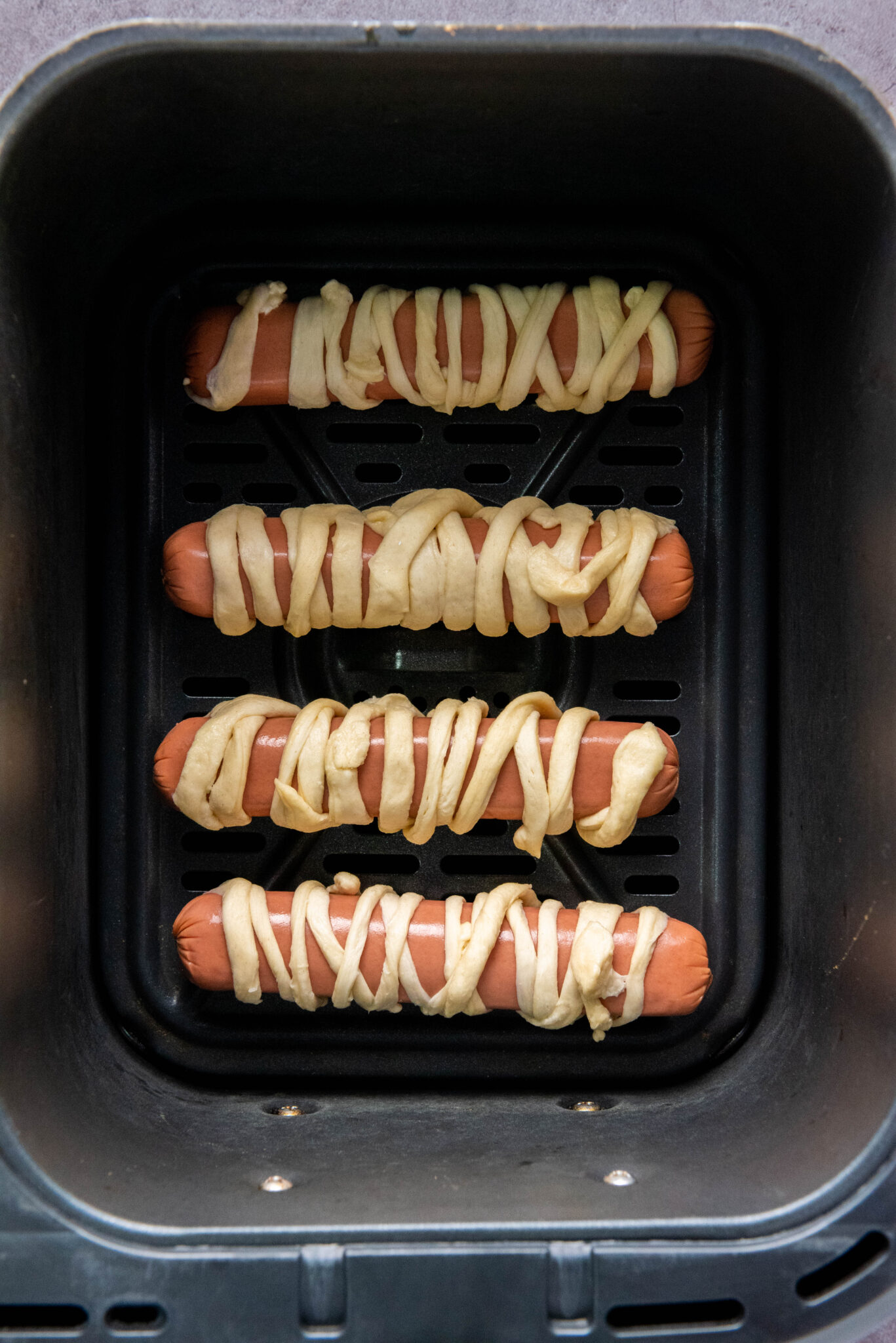 Four hot dogs wrapped in dough are placed in an air fryer basket, ready to be auto-drafted into a delicious meal.