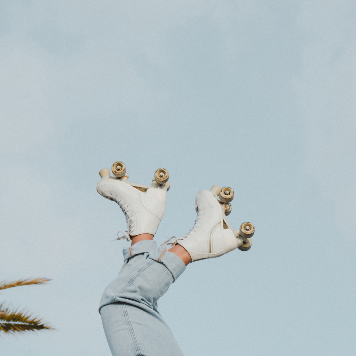 A person wearing light blue jeans and white roller skates with legs pointed upwards against a clear blue sky. Can you roller skate outside? A small part of a palm tree is visible in the lower left corner.