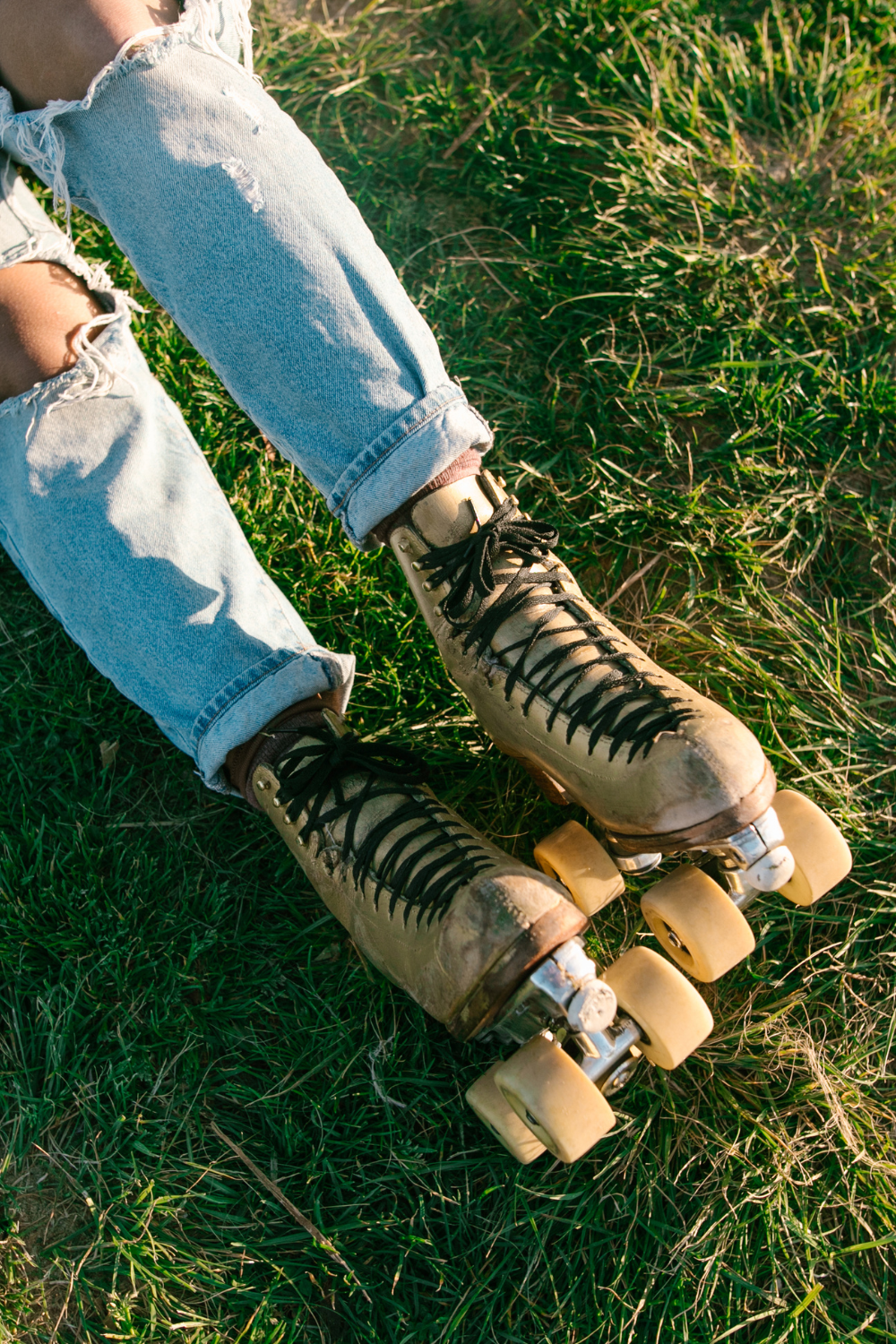 A person wearing worn roller skates and ripped jeans is sitting on the grass, pondering, "Can you roller skate outside?