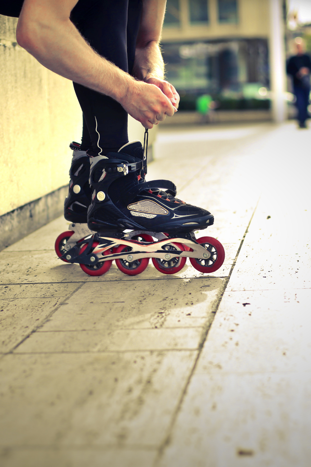 Person wearing rollerblades kneels to secure the laces, preparing to skate on a paved outdoor path. Can you roller skate outside and feel the wind in your hair?