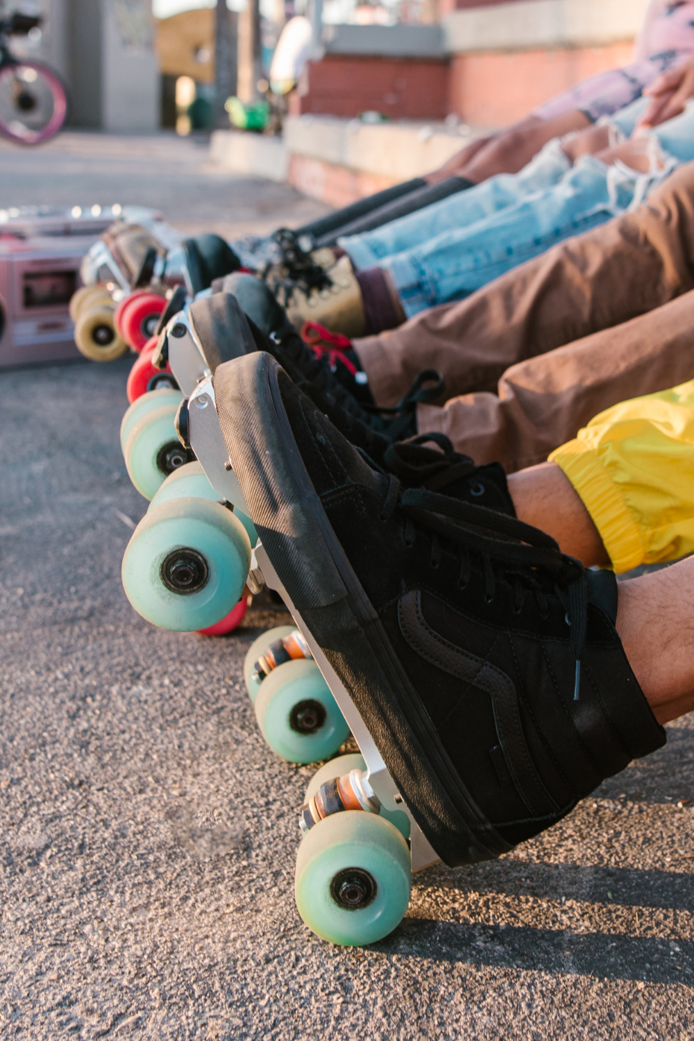 A row of people wearing roller skates, sitting on the ground. They all sport black sneakers with colorful wheels, as if pondering, "Can you roller skate outside?