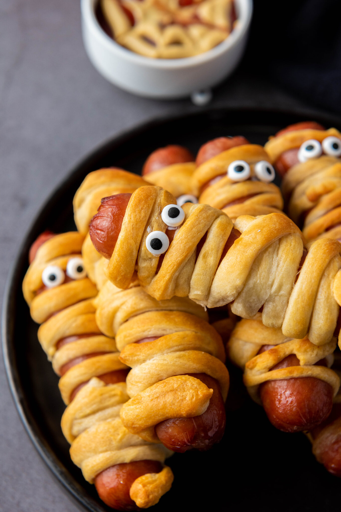 A plate of Air Fryer Halloween Mummy Dogs wrapped in dough to resemble mummies with edible eyes, accompanied by a bowl of crescent roll dip.