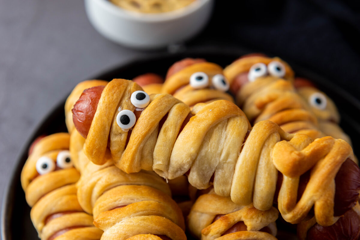 Close-up of hot dog mummies wrapped in crescent dough with candy eyes on a black plate. A dipping sauce in a white bowl is visible in the background.