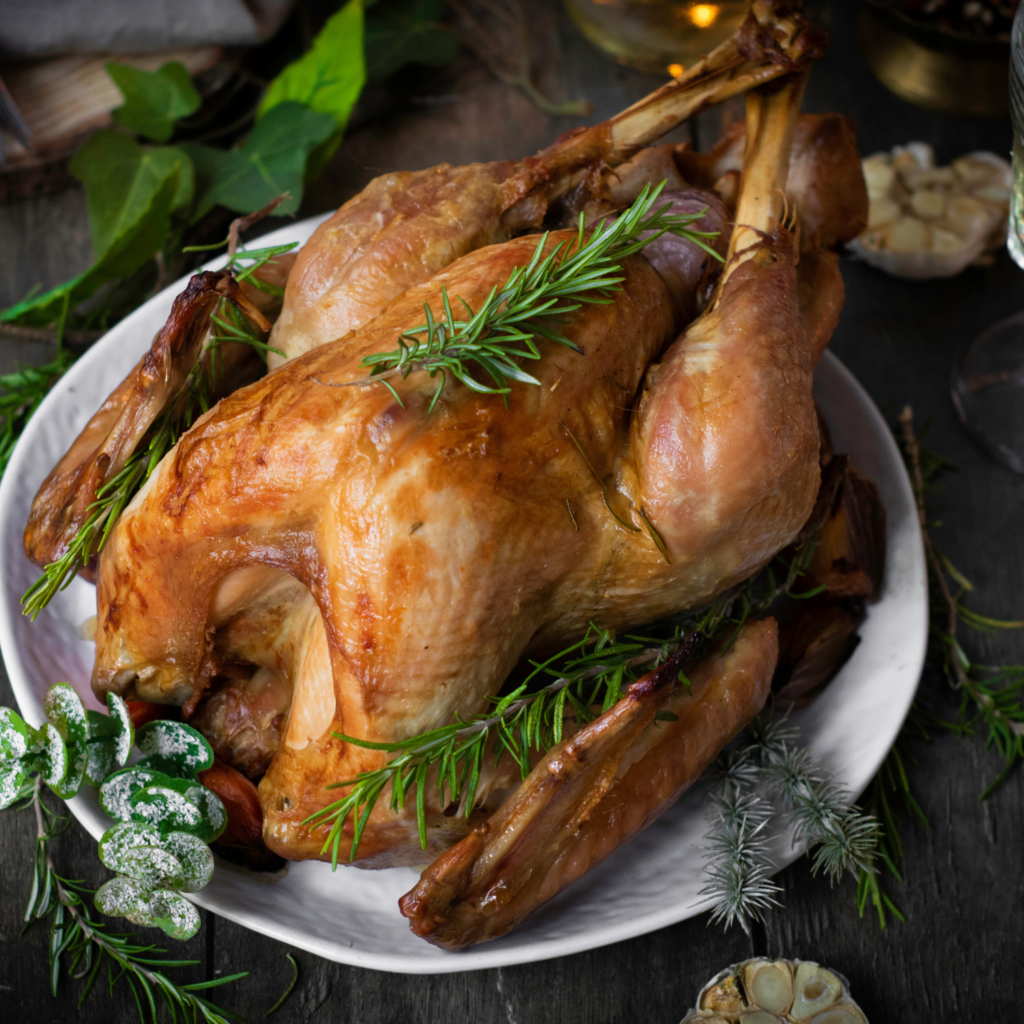A roasted Thanksgiving turkey garnished with fresh rosemary sits on a white plate, surrounded by various herbs and garlic on a rustic wooden table.