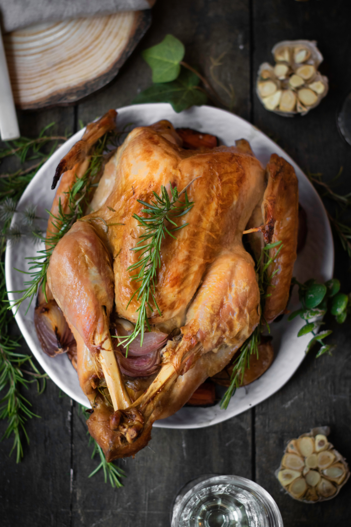 A roasted whole turkey garnished with fresh rosemary is placed on a white plate with roasted vegetables, evoking the comforting essence of a traditional Roasted Thanksgiving Turkey. Garlic and herbs are scattered around on a dark wooden table.