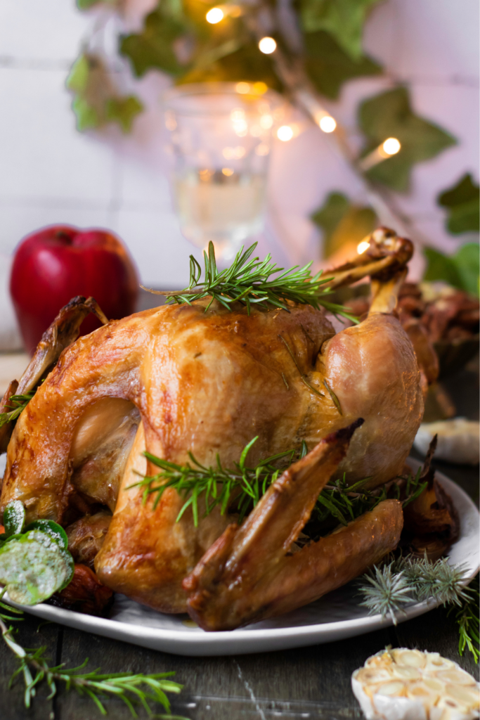 A roasted whole turkey garnished with sprigs of rosemary on a plate. An apple, garlic, and a glass are in the background along with festive lights and leaves, perfect for your next Thanksgiving Turkey Recipe inspiration.