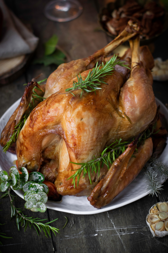 Roasted turkey garnished with sprigs of rosemary on a white platter, accompanied by sugared berries and roasted garlic, set on a rustic wooden table—an ideal Thanksgiving centerpiece.