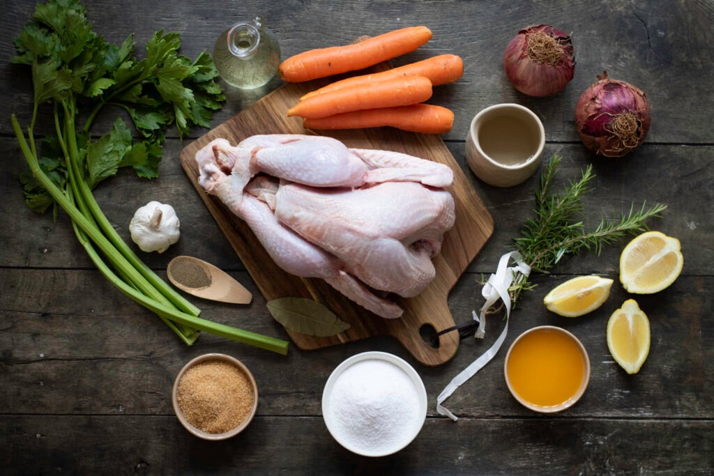 A wooden table holds raw chicken on a cutting board, surrounded by cooking ingredients: celery, green onions, garlic, spices, olive oil, carrots, red onions, rosemary, lemon, and bowls of sugar. This setup could easily become the starting point for a delicious roasted turkey recipe.