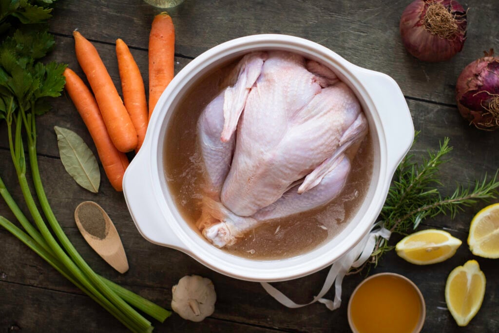 A whole raw chicken in a pot of water, surrounded by carrots, celery, garlic, onions, rosemary, and lemon wedges hints at the essence of a best turkey recipe for Thanksgiving. A small bowl of yellow liquid sits on a wooden surface.