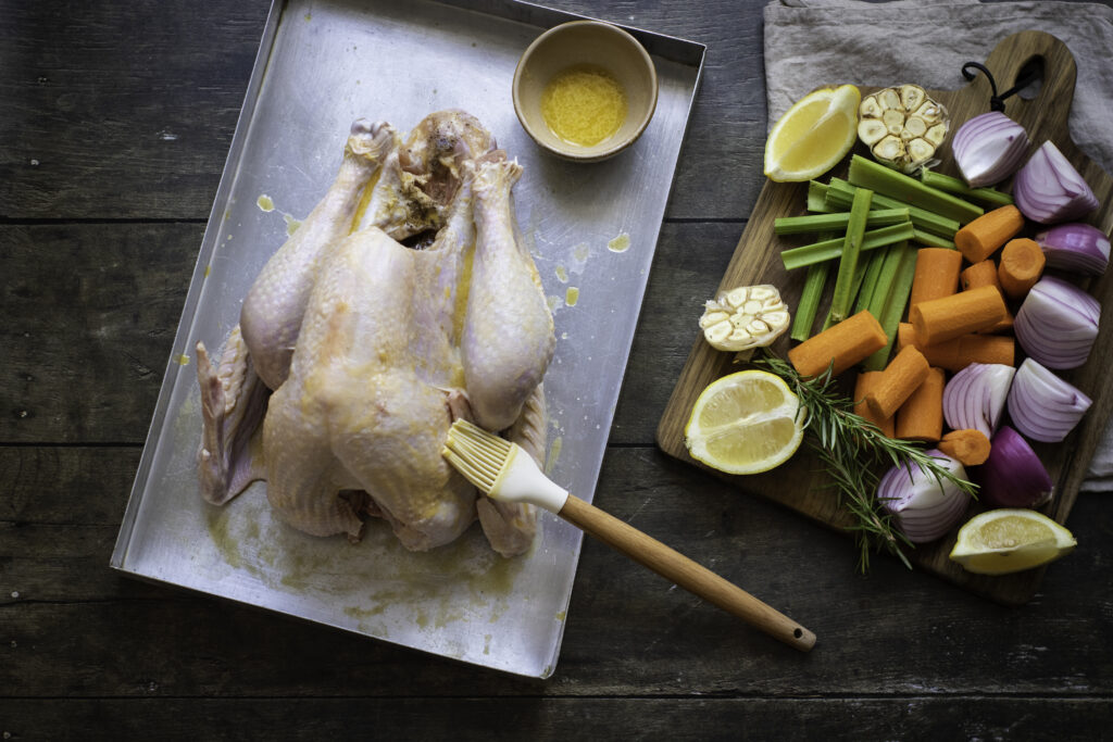 A raw chicken being brushed with seasoning sits on a baking sheet next to a cutting board with chopped vegetables, including carrots, onions, celery, garlic, and lemon—setting the stage for a roasted Thanksgiving turkey recipe.