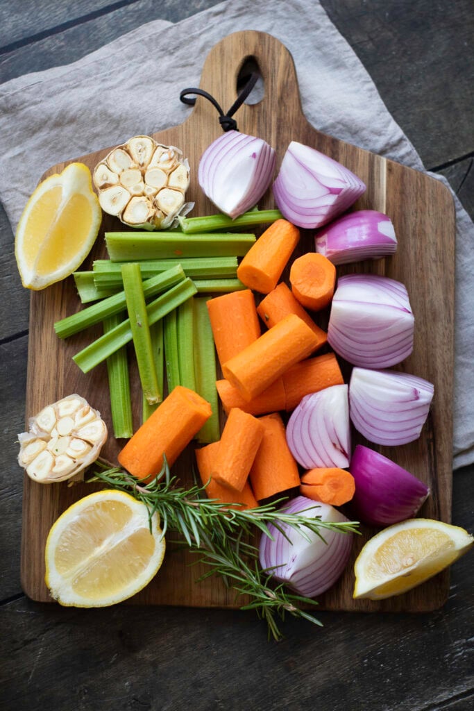 A wooden cutting board with chopped red onions, carrots, celery, halved garlic bulbs, lemon wedges, and a sprig of rosemary on a gray cloth background – the perfect prep for your Best Turkey Recipe this Thanksgiving.