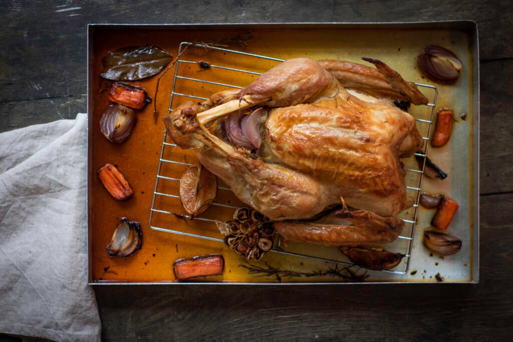 A roasted Thanksgiving turkey sits on a rack in a baking tray, surrounded by roasted vegetables and herbs. A beige cloth is partially visible on the side.