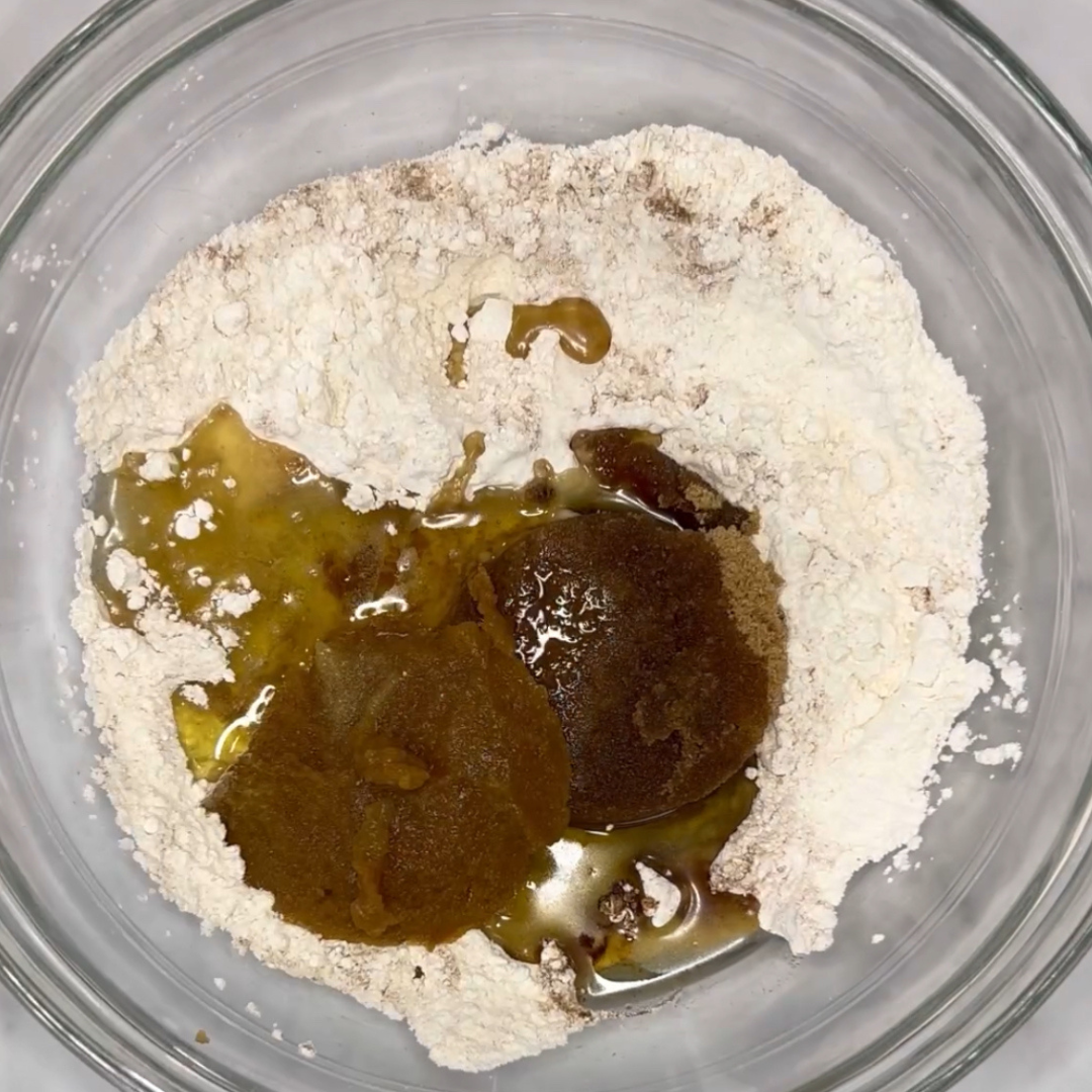 A glass bowl containing flour, brown sugar, granulated sugar, and liquid ingredients ready to be mixed for an easy one-bowl peach muffin recipe.