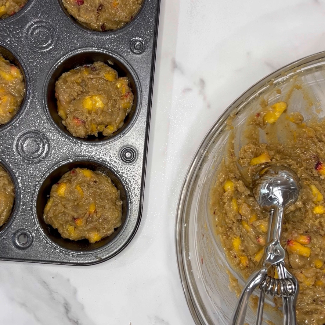 A muffin tin with batter in the cups sits on a countertop next to a glass bowl containing more batter and an ice cream scoop, perfect for making One Bowl Peach Muffins.