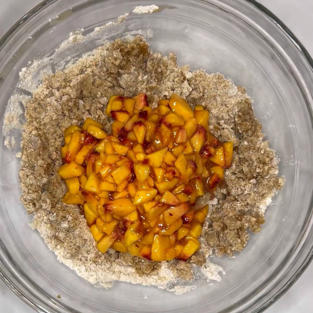A glass mixing bowl containing chopped peaches in the center is surrounded by a light brown mixture, likely of flour and other dry ingredients, perfect for an easy one-bowl peach muffin recipe.
