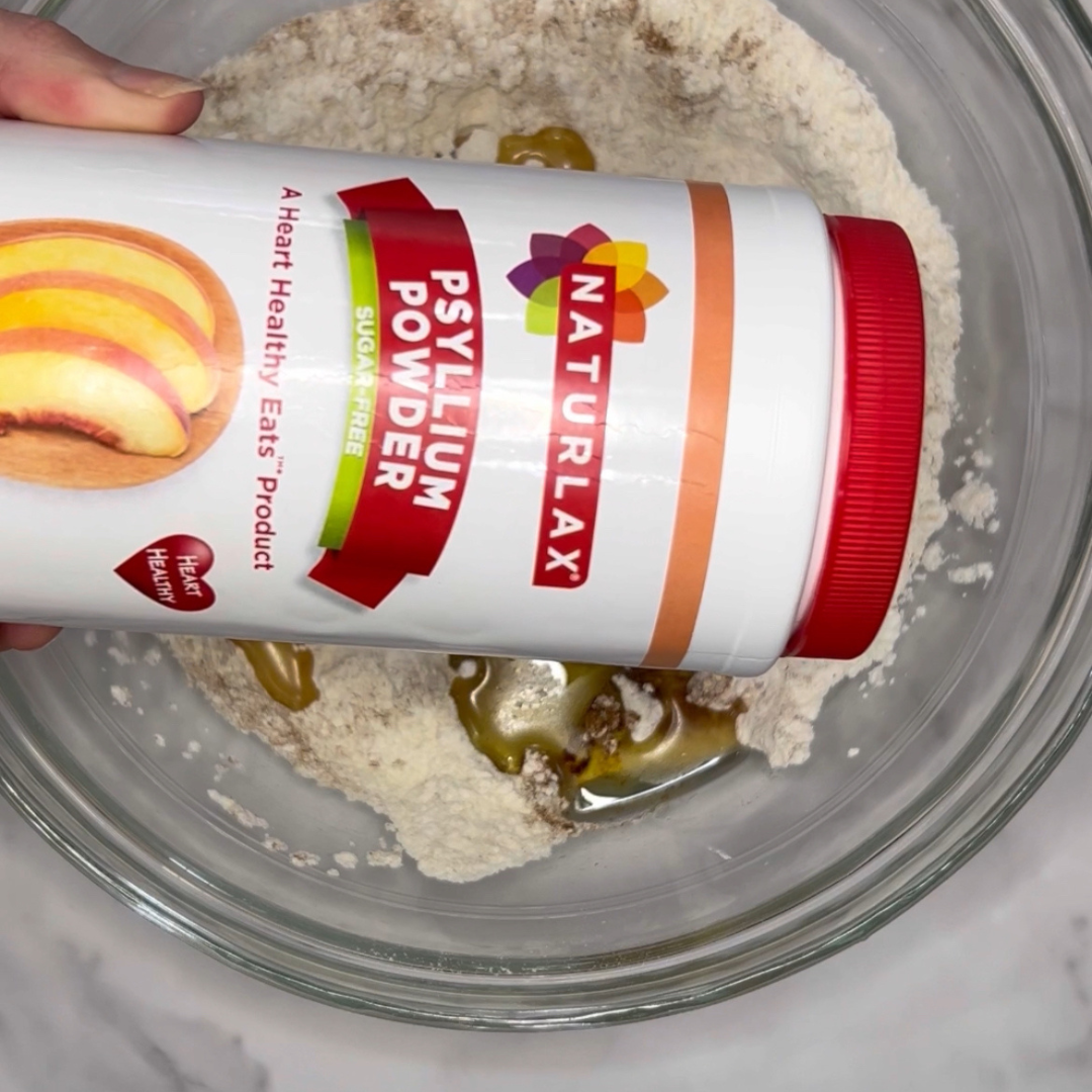 A person is pouring Naturalax Psyllium Powder from a container into a glass bowl with dry ingredients for an Easy Peach Muffins recipe.