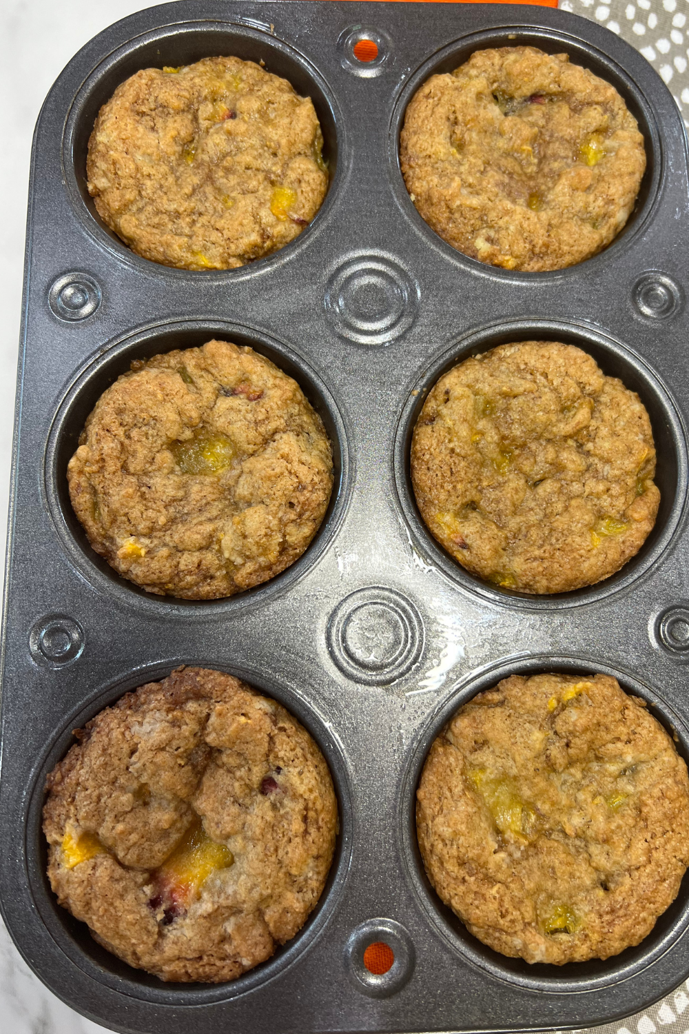 A tray of six freshly baked, easy one-bowl peach muffins with visible pieces of fruit sits invitingly on a kitchen counter.